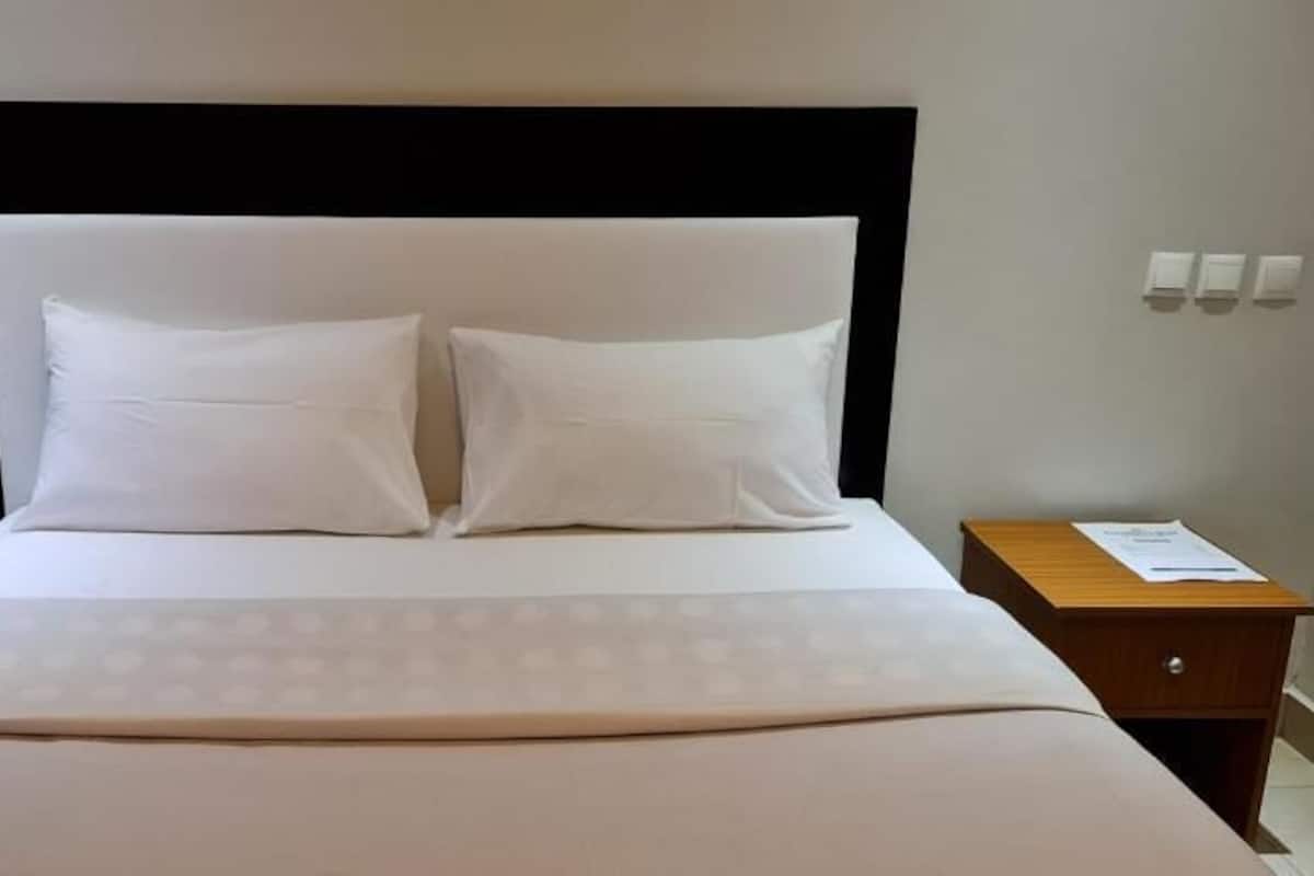 A neatly made bed with a white headboard and pillows, a wooden nightstand with a lamp, and a small table with a menu or information card.