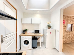 A compact kitchen with a washing machine, a microwave, a small refrigerator, and a wooden desk with a plant.