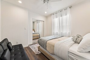 Un dormitorio con cama, sofá negro, ventilador de techo y vista a otra habitación a través de un espejo.