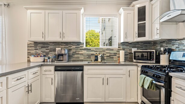 A modern kitchen with white cabinets, stainless steel appliances, a tiled backsplash, and a window with blinds.