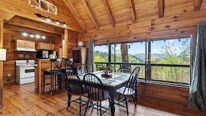 A wooden cabin interior with a dining area, kitchen, and large windows offering a view of the outdoors. - Sleeps 42, 7 Cabins Private Hot Tubs + Shared Fire Pit + Mountain Views + Covered Decks 7 Min to Townsend (Townsend)