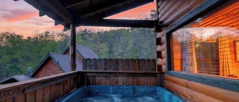 A wooden cabin with a hot tub, a wooden deck, and a view of the forest.