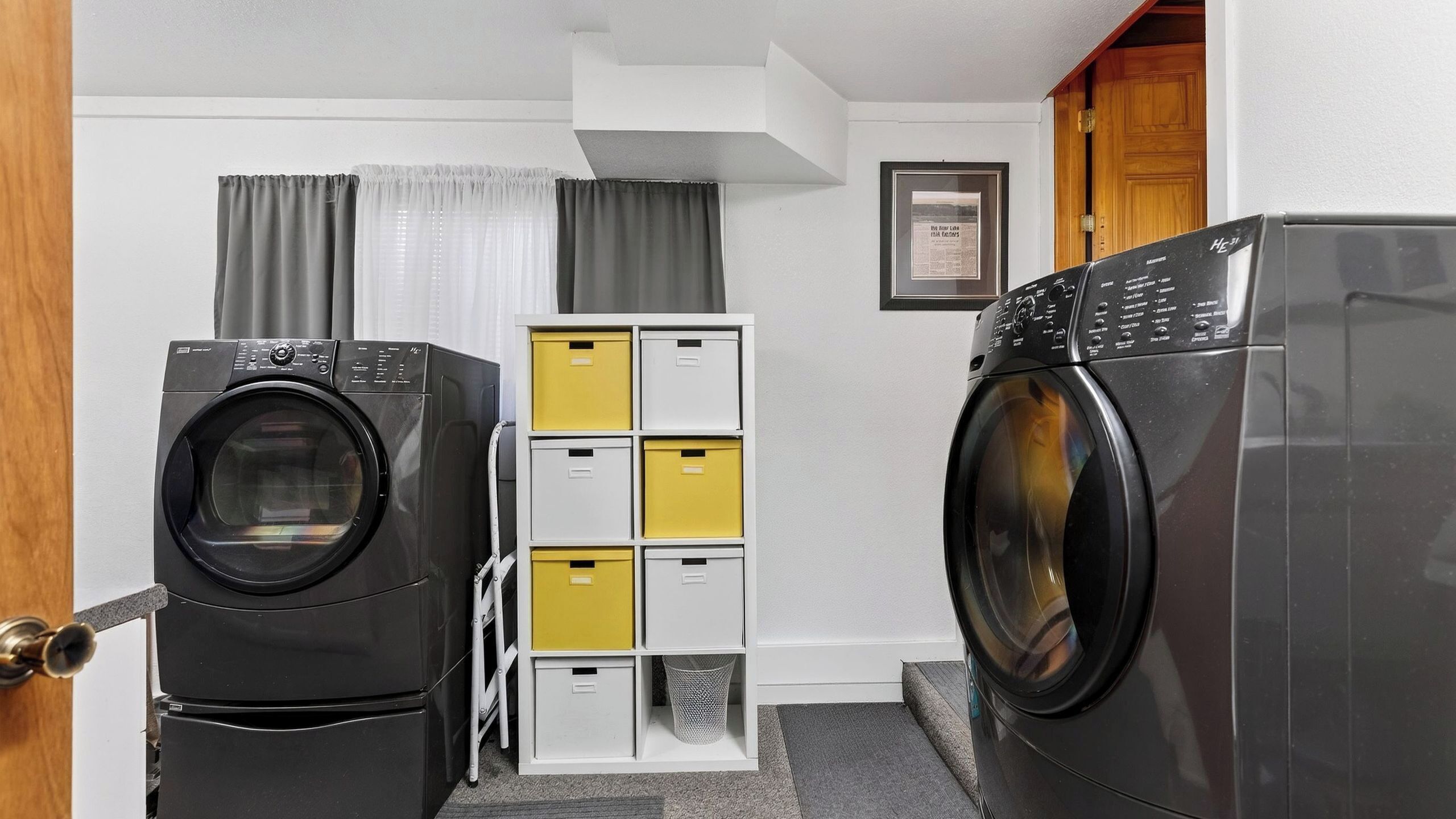 Une buanderie équipée de deux lave-linge et deux sèche-linge à chargement frontal, d’une étagère métallique avec des bacs de rangement jaunes et blancs, et d’un certificat encadré accroché au mur.