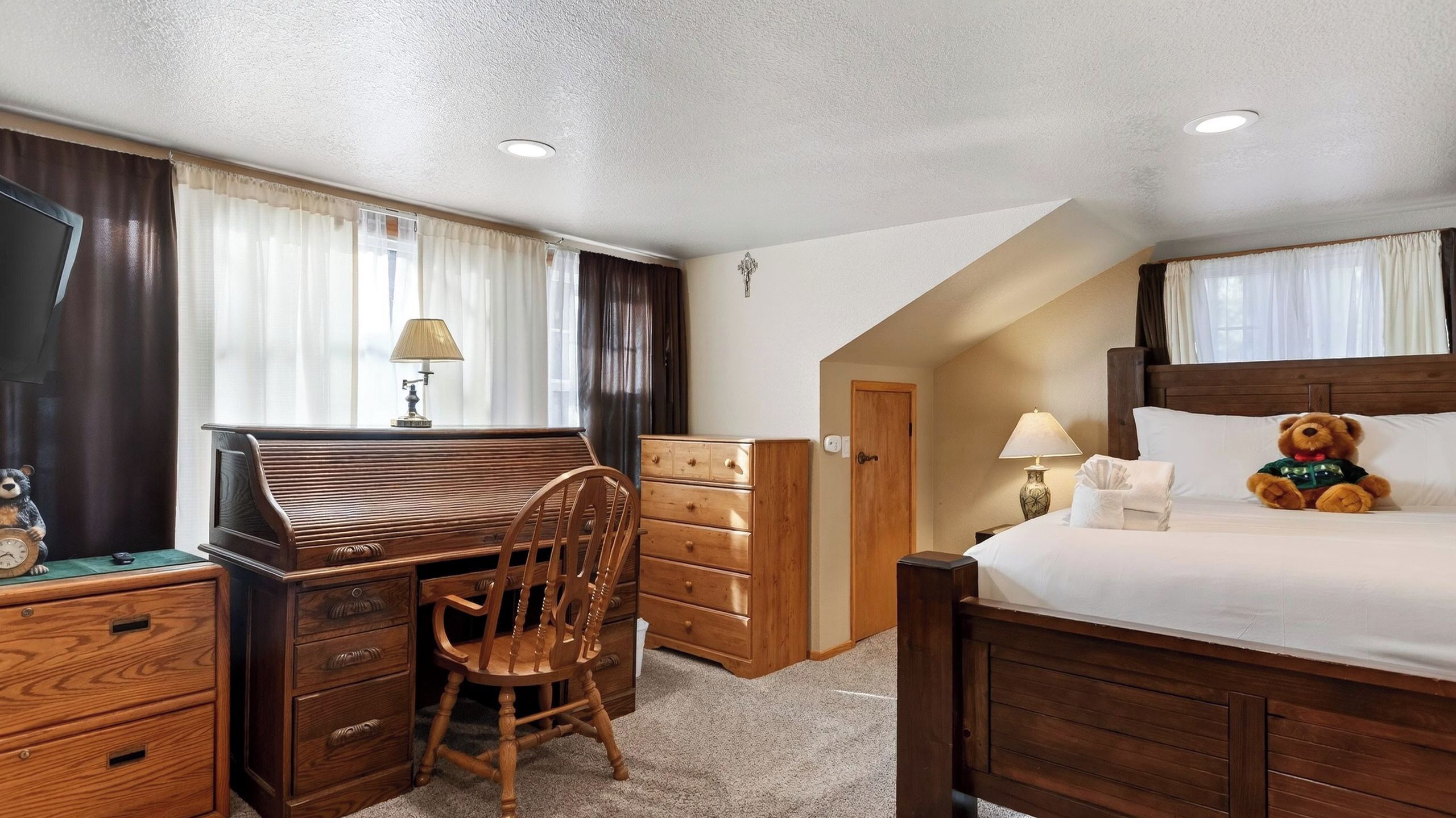 A bedroom with a wooden desk, chair, dresser, and a bed with a teddy bear.