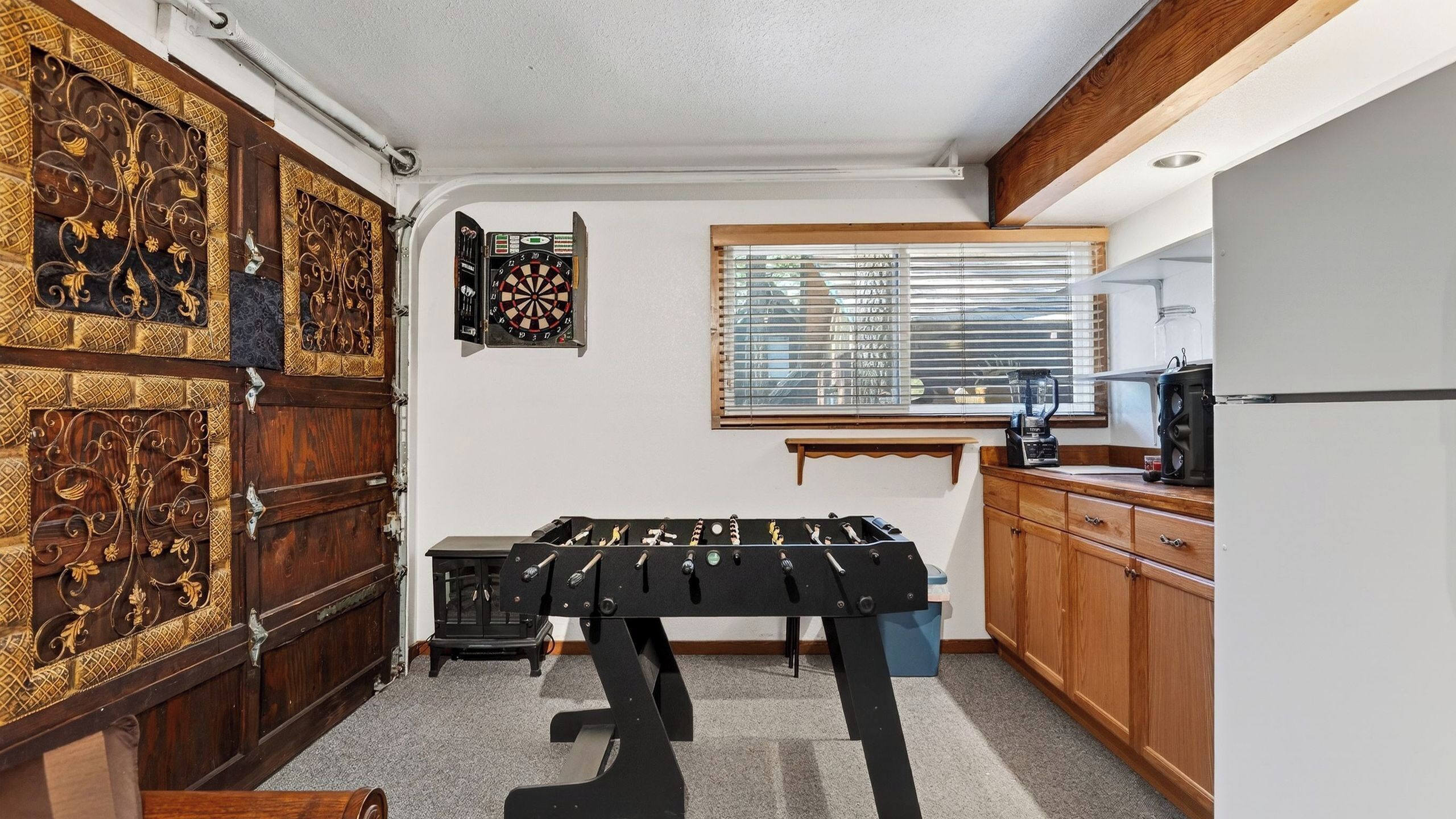 A compact kitchen with a dartboard, a window with blinds, and a coffee machine.