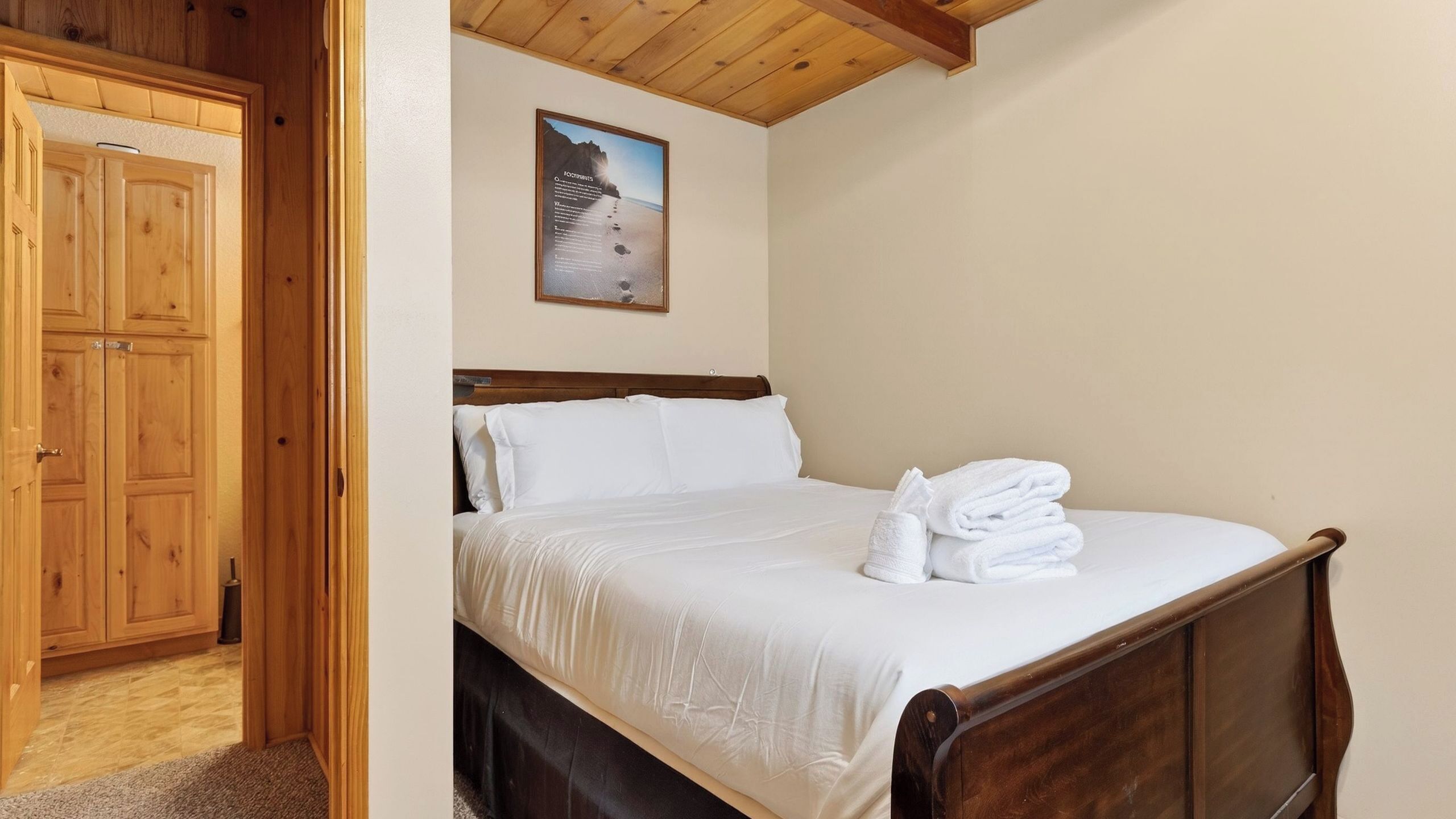A bedroom with a wooden bed frame, white bedding, a wooden wardrobe, and a framed picture on the wall.