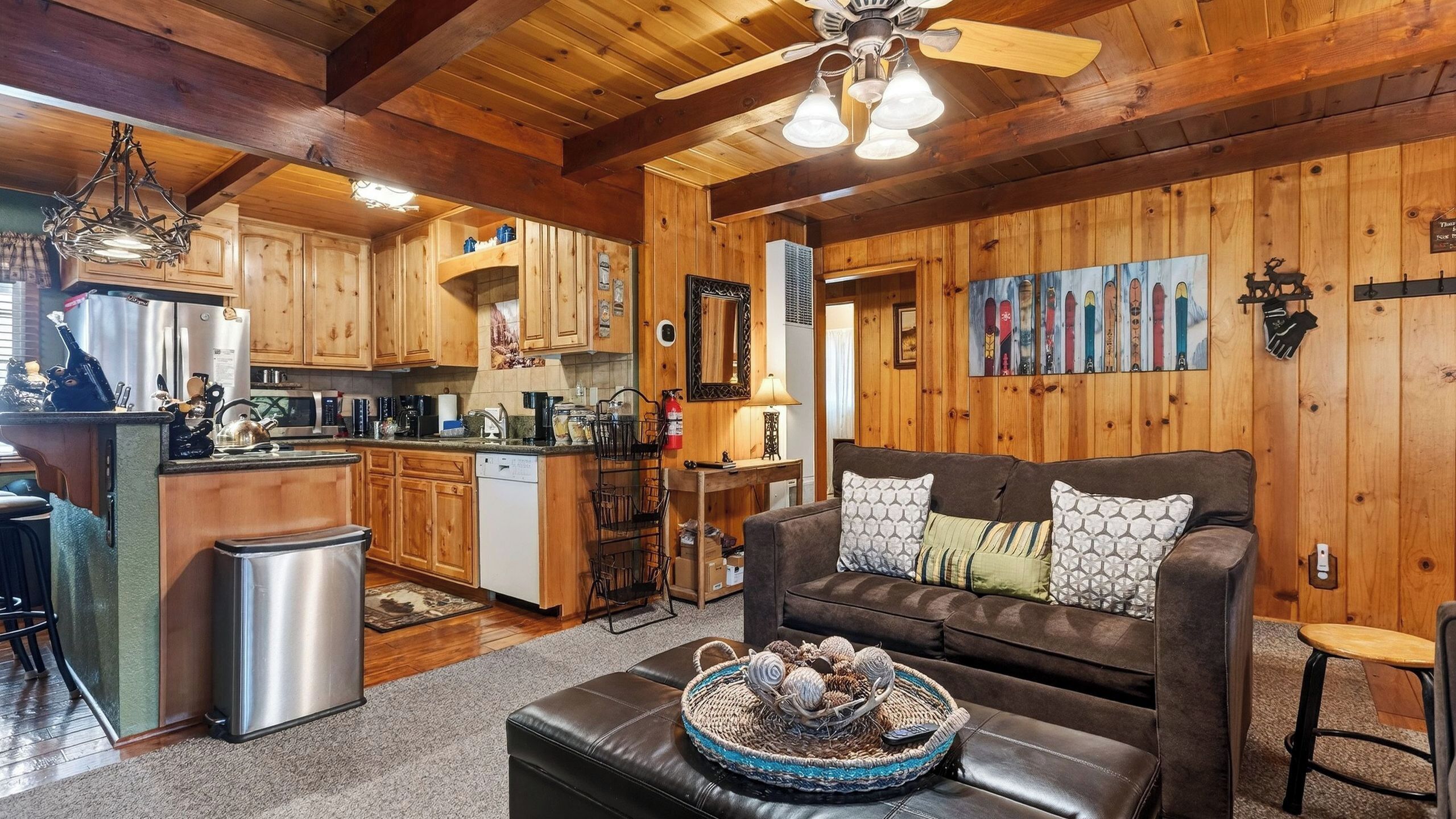 A cozy cabin interior with wooden walls, a kitchen area, a living room with a sofa and decorative pillows, and a ceiling fan.