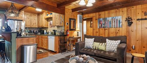A cozy cabin interior with wooden walls, a kitchen area, a living room with a sofa and decorative pillows, and a ceiling fan.