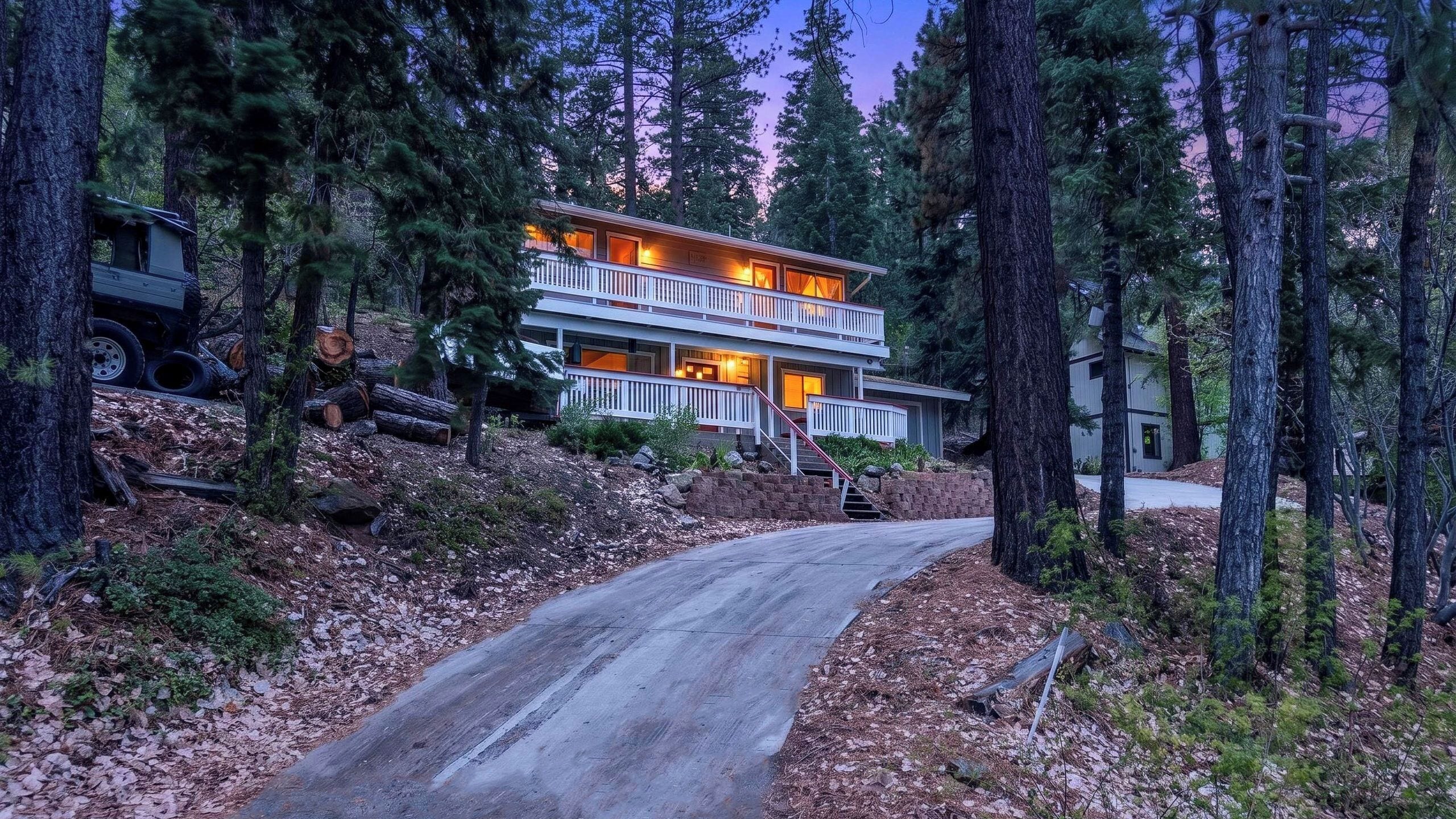 A two-story cabin with a balcony, surrounded by trees and a driveway.
