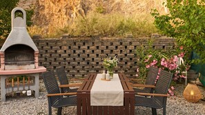 Een terras buiten, met een bakstenen muur, een stenen barbecue, een tafel met een tafelkleed en twee stoelen.