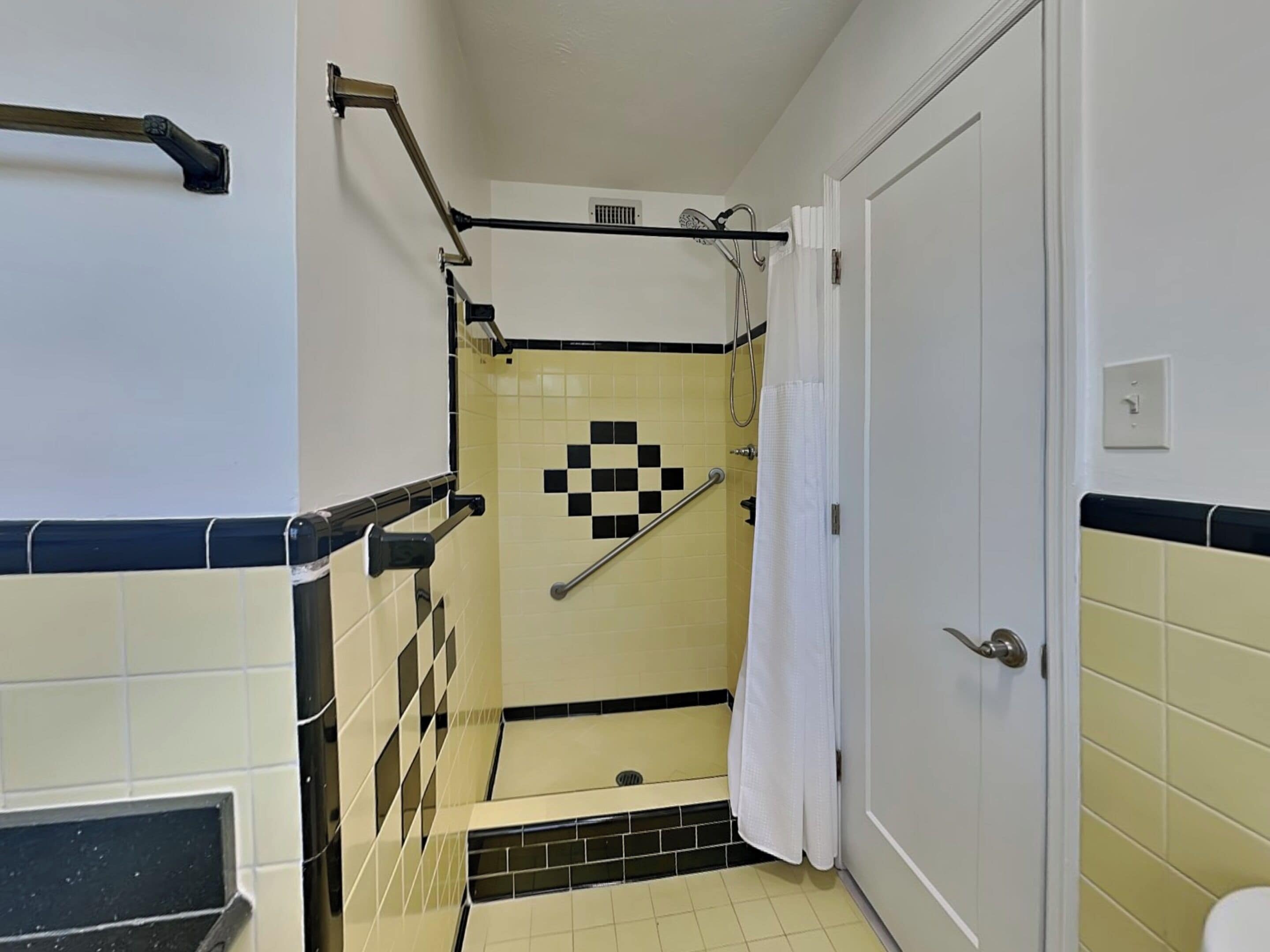 Une salle de bain avec un carrelage à motifs géométriques, une douche équipée d’une barre d’appui et une porte blanche.
