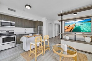 Eine moderne Küche mit Mikrowelle, Backofen und Kühlschrank, ein Essbereich mit Stühlen und Tisch sowie ein Fernseher, der eine tropische Szene zeigt.