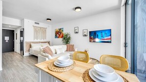 Een moderne woonkamer met een bank, eettafel en stoelen. Er is een groot raam, een plant en kunst aan de wanden.