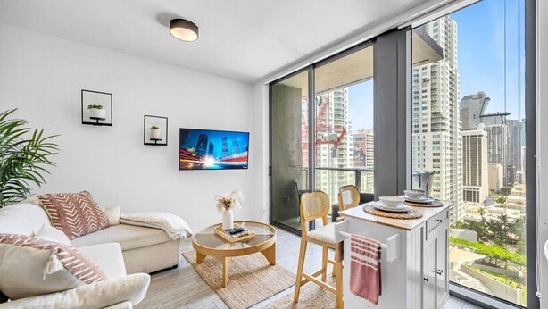Een moderne woonkamer met een bank, een salontafel en een eethoek met stoelen. Een groot raam biedt uitzicht op de stad.