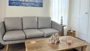 Una sala de estar con un sofá gris, una mesa de centro de madera, un cuadro de veleros, una puerta blanca y una lámpara.