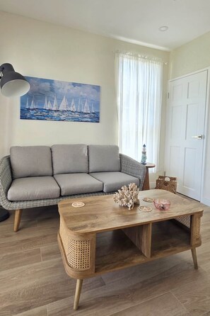 A living room with a grey sofa, a wooden coffee table, a painting of sailboats, a white door, and a lamp. - Two BD Villa No.2, West Rock Villas, St James (Mount Standfast)