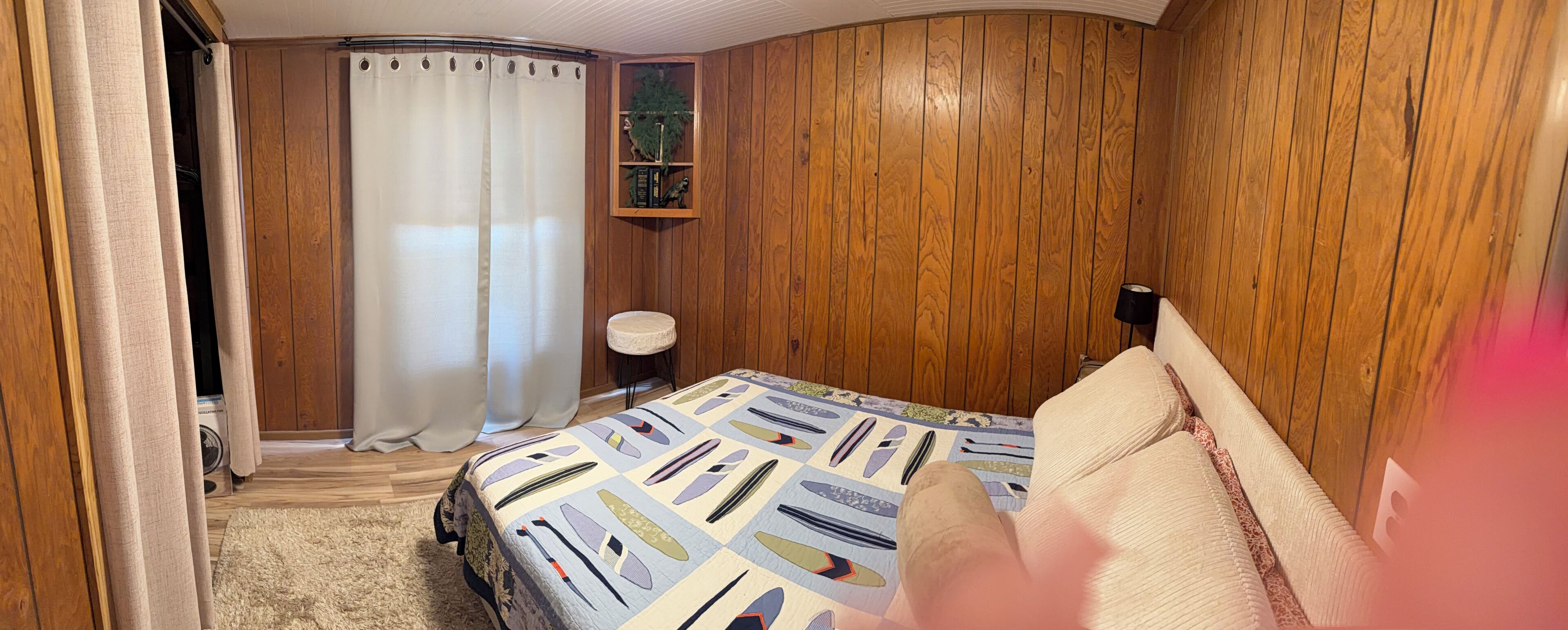 Un dormitorio con pared de paneles de madera, una cama con colcha estampada, un taburete pequeño y una estantería con libros y una planta.