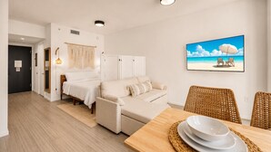 A room with a bed, a sofa, a dining table with plates, a wicker chair, and a beach scene painting on the wall. - Casa Soléa Apartment | Luxe Miami Escape (Miami)