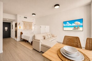 Quarto com cama, sofá, mesa de jantar com pratos, uma cadeira de vime e uma pintura de uma cena de praia na parede.