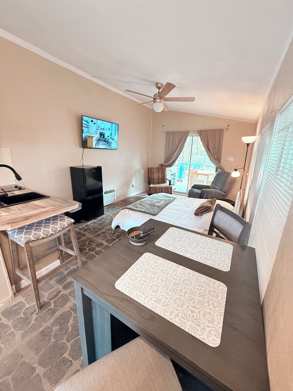 A hotel room with a dining area, a TV mounted on the wall, a ceiling fan, and a view of the outside.