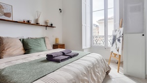 Un dormitorio con cama, almohadas, una colcha, una mesita de noche, un estante con objetos decorativos, un ventanal con vistas y un caballete con una pintura.