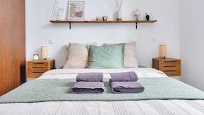 Una cama bien tendida con dos toallas dobladas, una lámpara de noche, una mesita de noche, una estantería con objetos decorativos y una obra de arte enmarcada en la pared.