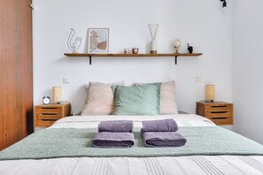 A neatly made bed with two folded towels, a bedside lamp, a nightstand, a shelf with decorative items, and a framed artwork on the wall. - Bright apartment NICE CENTRE - 4 pers - Near Beach & Place Masséna (Nice)
