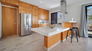 A modern kitchen with wooden cabinets, a stainless steel refrigerator, a central island with stools, and a large window with curtains. - Stunning & Beautiful House With Private Pool (Cabo San Lucas)