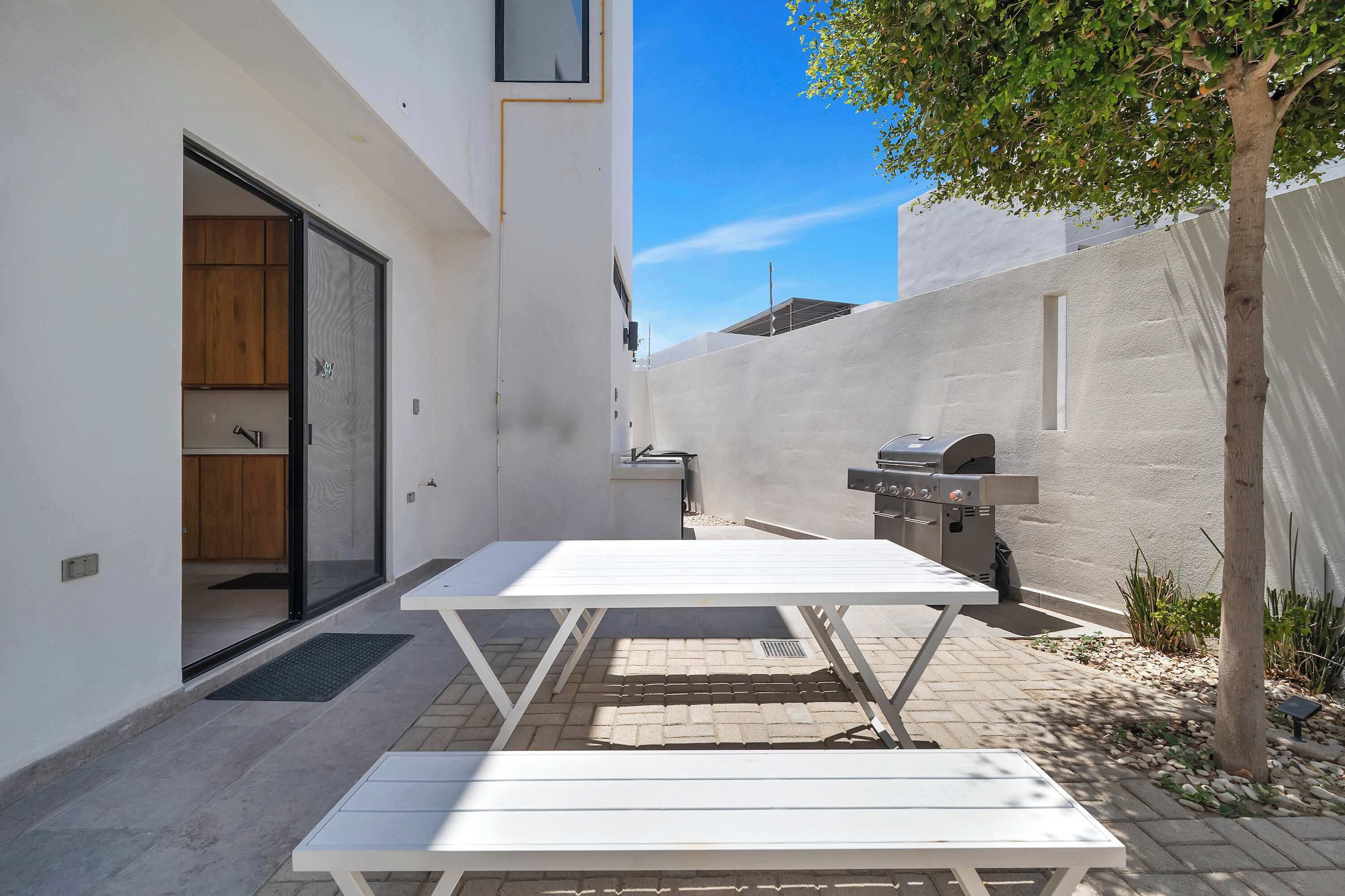 Un patio con una mesa, bancos, una puerta corrediza, una parrilla y un árbol.