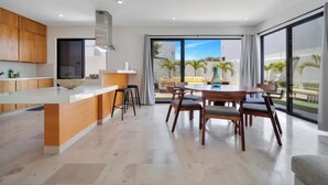 A modern kitchen with a central island, a dining area with wooden chairs, and large sliding glass doors leading to a balcony with palm trees. - Stunning & Beautiful House With Private Pool (Cabo San Lucas)