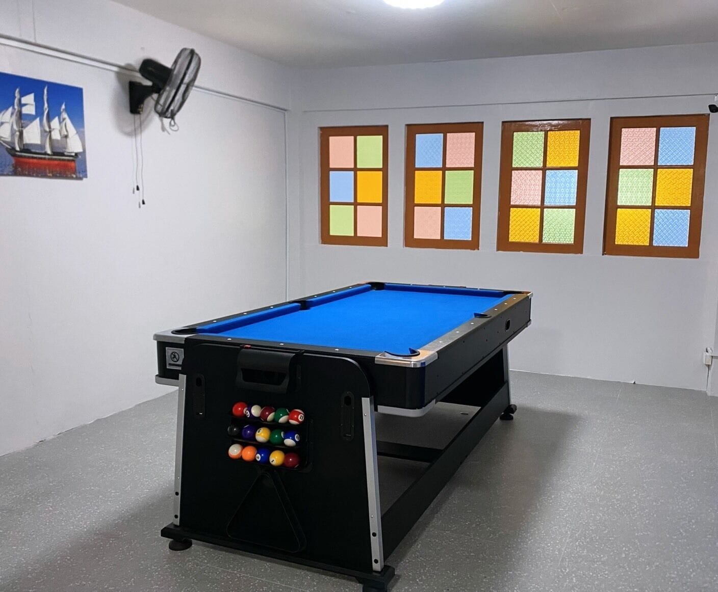 A pool table with a blue felt surface, a rack of billiard balls, and four windows with colorful panes.