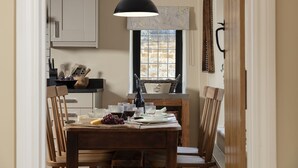 A dining area with a wooden table, chairs, a hanging light fixture, a window with a view, and kitchen utensils on the counter. - Sun Cottage - Wonderful dog friendly cottage (Kingham)