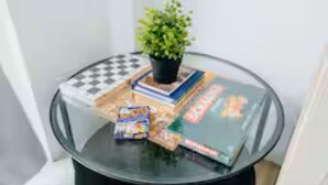 Una mesa con cubierta de vidrio, una planta en maceta, un tablero de ajedrez, algunos libros y un paquete de comida.