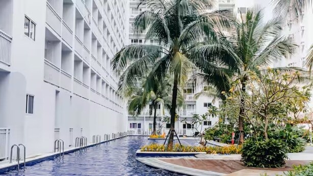 Una piscina con un edificio blanco al fondo, palmeras y una zona ajardinada bien cuidada.