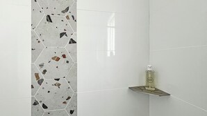 A bathroom with white tiles, a hexagonal mosaic pattern on the wall, a small shelf with a bottle, and a shower control panel.