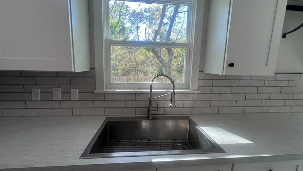 A kitchen with a stainless steel sink, white cabinets, a window with a view of trees, and a white tiled backsplash. - Beautiful 2-bedroom house with AC in peaceful Palm Valley (Palm Valley)