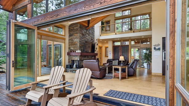 Ein modernes Haus mit Kamin aus Stein, einer Holzterrasse und Glastüren, die in einen Wohnbereich mit Möbeln führen.
