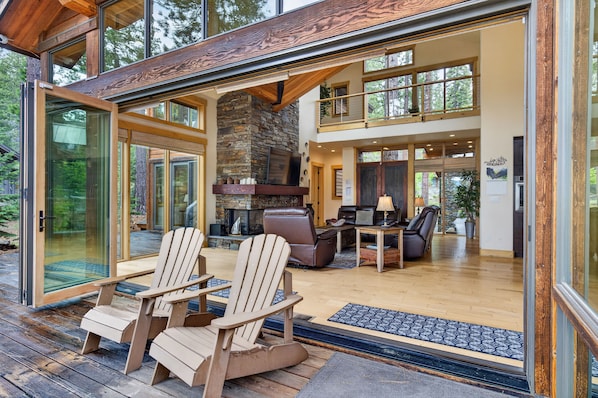 Ein modernes Haus mit Kamin aus Stein, einer Holzterrasse und Glastüren, die in einen Wohnbereich mit Möbeln führen.