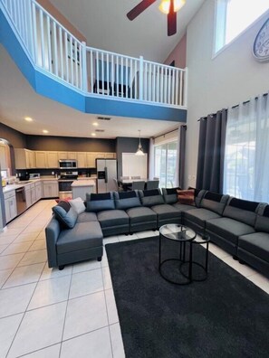 A spacious living area with a large sectional sofa, a small round coffee table, and a kitchen in the background. - Emerald Island Resort Home 6bdrm, Pvt Large Pool-10min to Disney! (Kissimmee)