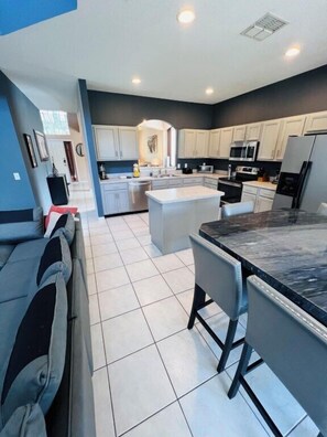 A modern kitchen with white cabinets, a central island, and a dining area with bar stools. - Emerald Island Resort Home 6bdrm, Pvt Large Pool-10min to Disney! (Kissimmee)