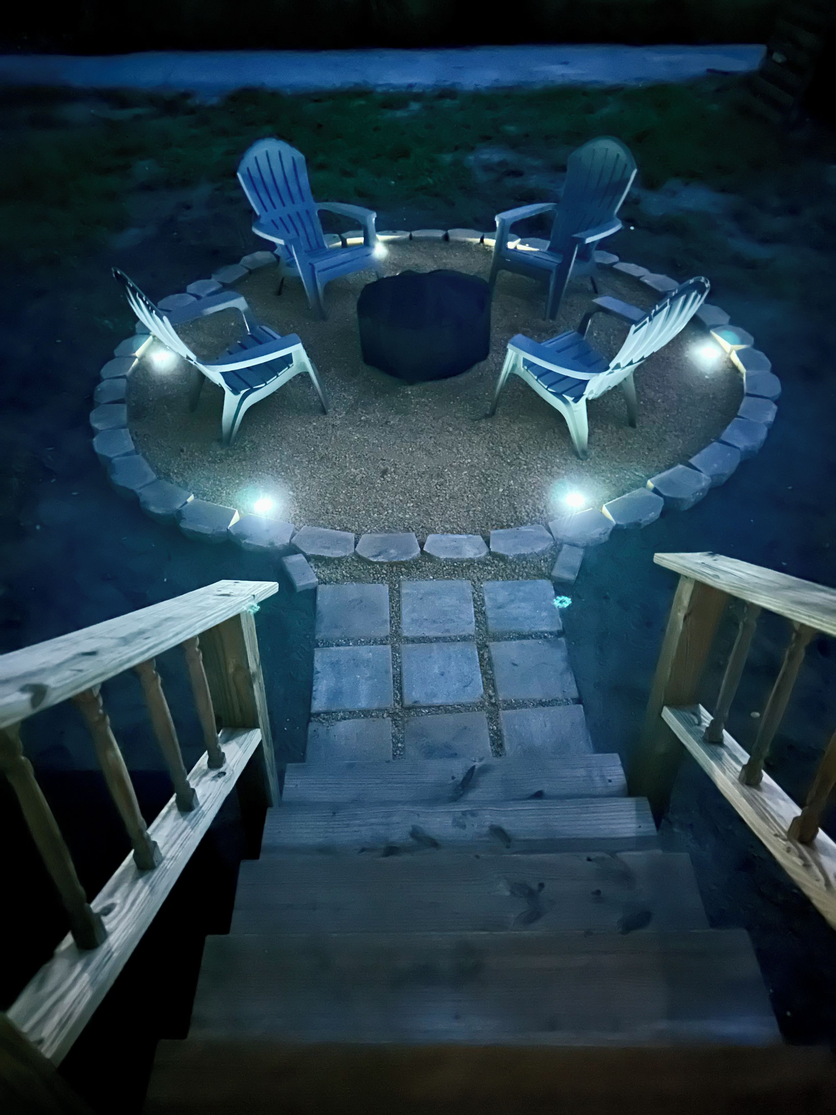 Un espacio de descanso circular al aire libre con sillas y un brasero, rodeado por un borde de piedra e iluminado con luces.