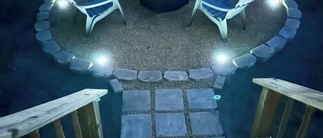 Un espacio de descanso circular al aire libre con sillas y un brasero, rodeado por un borde de piedra e iluminado con luces.