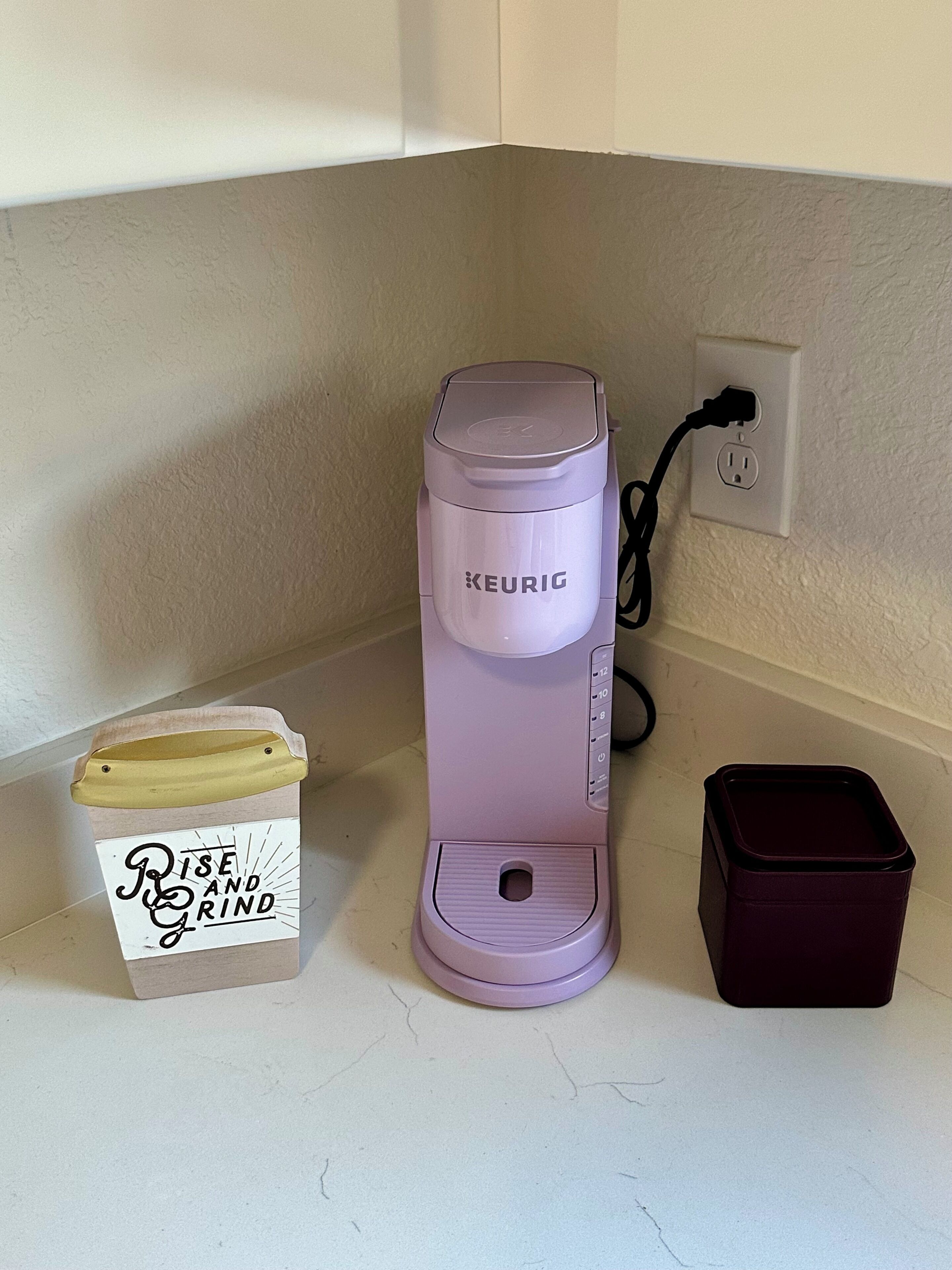 Een Keurig koffiezetapparaat op een aanrecht, een kleine prullenbak en een bakje met een motiverende quote.