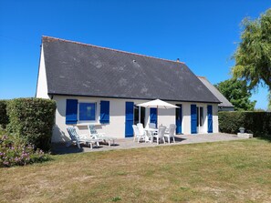 A two-story house with a tiled roof, a covered patio with white chairs and a table, and a garden area with grass and shrubs. - Holiday Home in Santec on Dossen Beach (Santec)