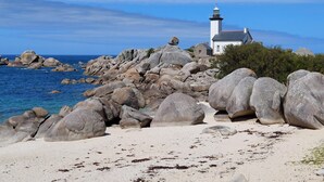 Ett fyrtorn på en klippig strand med klart, blått vatten och en vit byggnad i närheten.