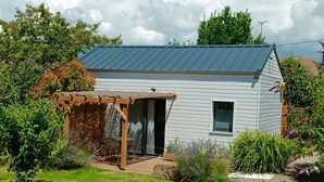 A small, single-story house with a metal roof, a wooden pergola, and a garden with various plants and flowers.