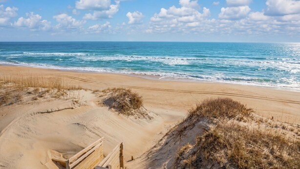 Une plage avec des dunes de sable, une promenade en bois et l’océan en arrière-plan.