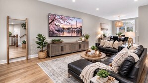 A modern living room with a large flat-screen TV, a leather sofa, a coffee table, and a dining area with a lamp.