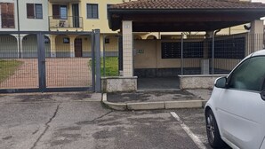 Een meerlaags woongebouw met een hekpoort, een overdekte parkeerplaats en een witte auto in de buurt.