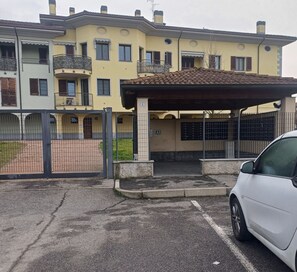 Ein mehrgeschossiges Wohngebäude mit geschlossenem Eingangsbereich, einem überdachten Parkplatz und einem weißen Auto in der Nähe.